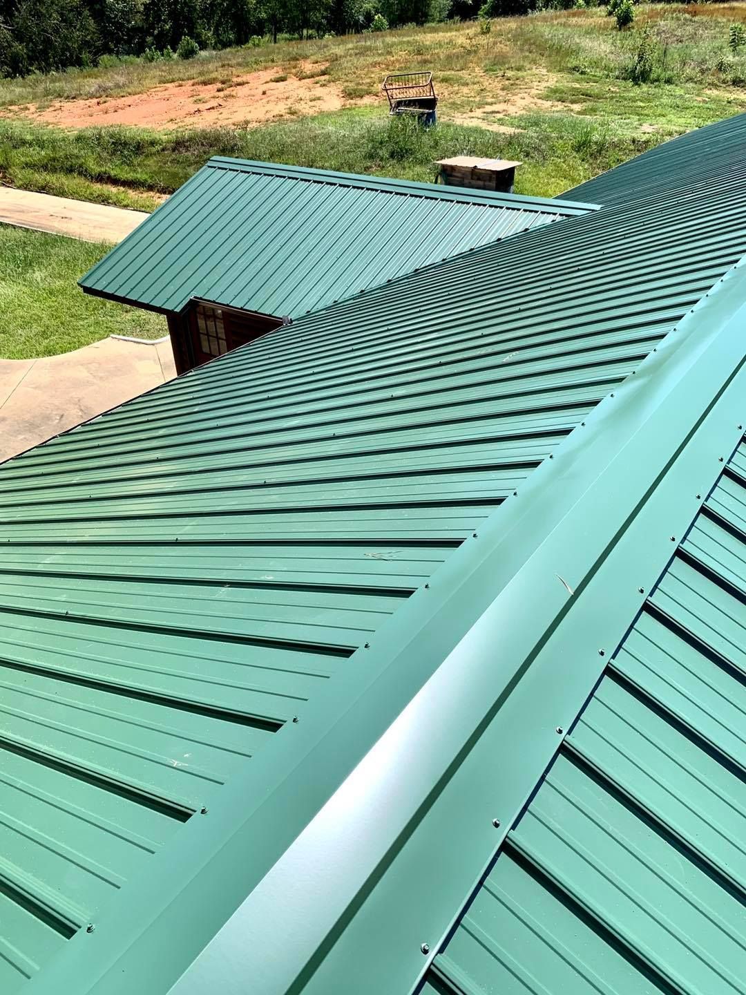 A high-angle view of a green metal roof with raised seams, featuring a ridgeline and a smaller intersecting roof section.