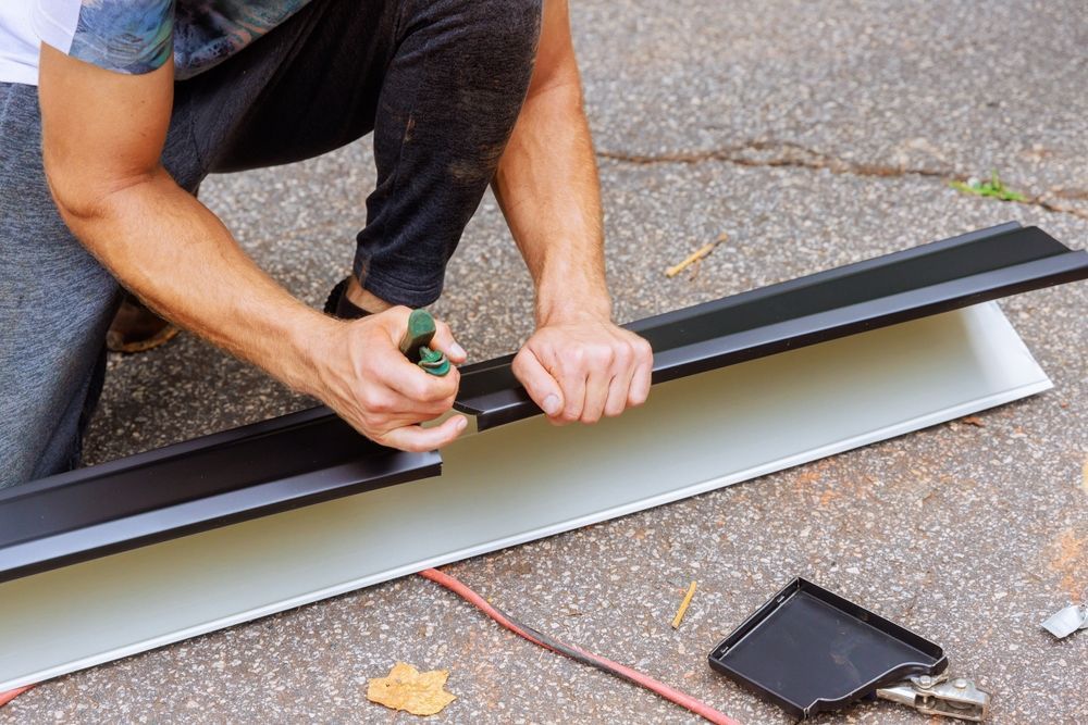 A person kneeling on pavement, using green metal snips to trim a piece of black and silver flashing.